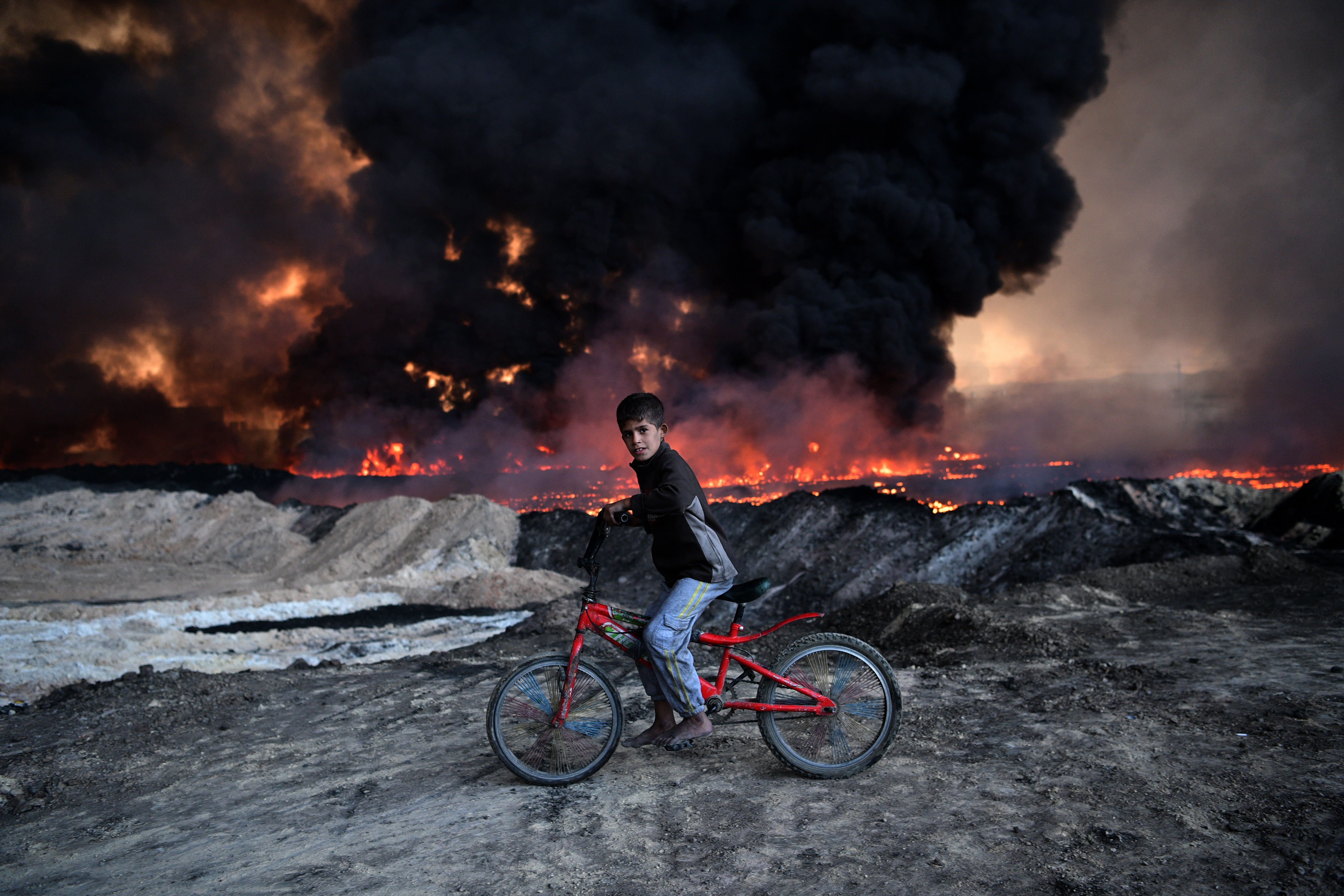 Un baiat pe bicicleta se opreste langa un camp petrolier incendiat de ISIS, in Mosul